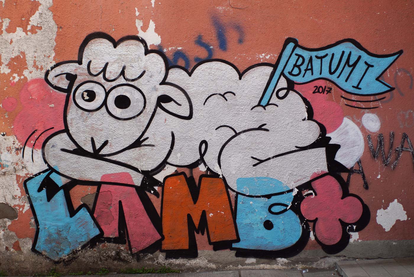 Zu sehen ist ein Graffiti Schaf das auf dem Schriftzug Lamb liegt. In dem hinteren Ende seines Felles steckt eine blaue Fahne mit der Aufschrift Batumi.