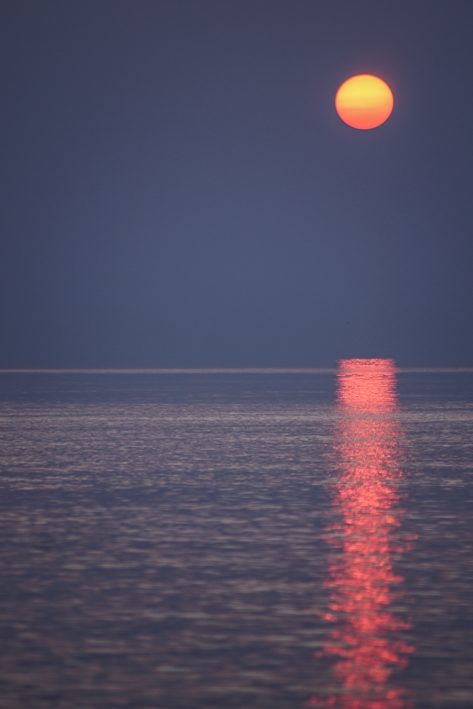 Zu sehen ist, wie die Sonne über einem spiegelglatten Meer untergeht,in diesem Spiegel sie sich orange rot wieder.