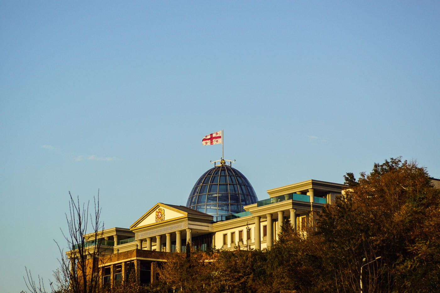 Zu sehen ist der Präsidentenpalast in Georgien Tbilisi. Auf seiner gläsernen Kuppel, ähnlich der des Reichstages, weht die georgische Flagge. Das ganze Gebäude ist in ein wunderschönes Licht durch die untergehende Sonne getaucht.