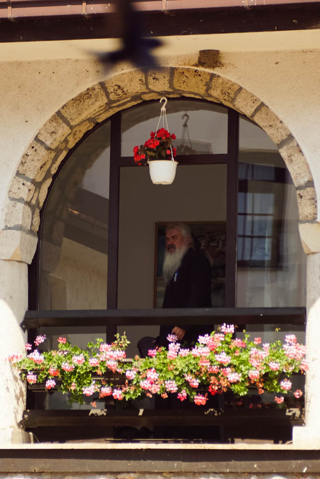 Zu sehen ist ein bogenförmiges Fenster aus dem ein Mönch mit langem weißem Bart schaut. Vor dem Fenster ist ein großer Blumenkasten mit Geranien angebracht von der Decke hängt noch ein Topf mit roten Blumen.