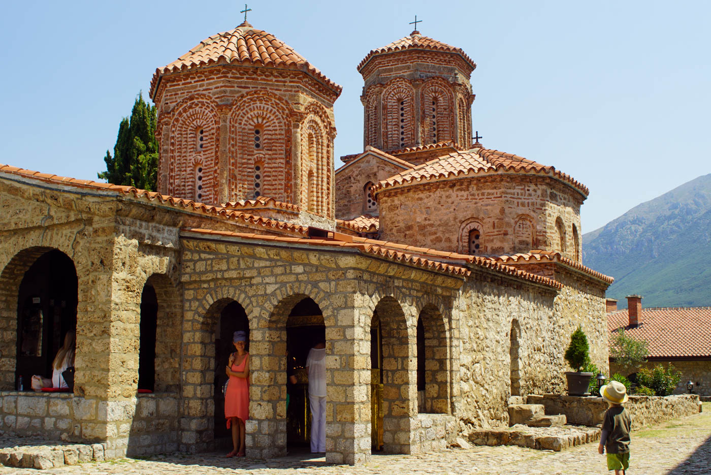 Im Vordergrund steht Luk mit Hut und blickt auf die Kirche von Sveti Naum. Lea steht in einem der Bögen und blickt lächelnd zu Luk