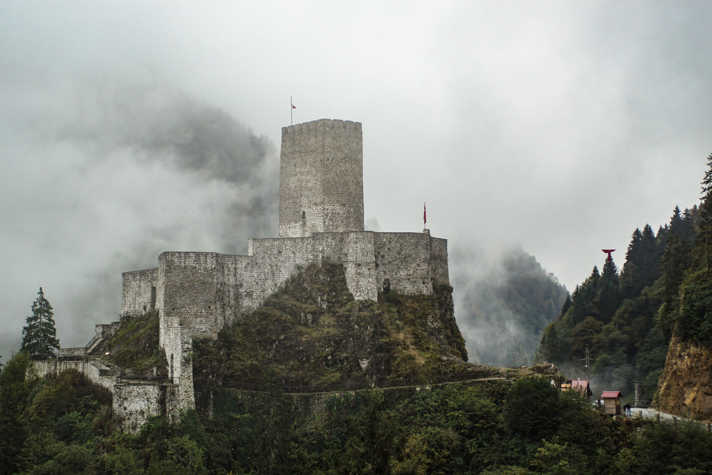 Zu sehen ist eine mittelalterliche Burg die sich auf einen Felsgrat festhält. Der Bergfried erhebt sich meterweise in die Höhe der Hintergrund ist wolkenverhangen und vereinzelt ist der gegenüberliegende Berghang zu erkennen.
