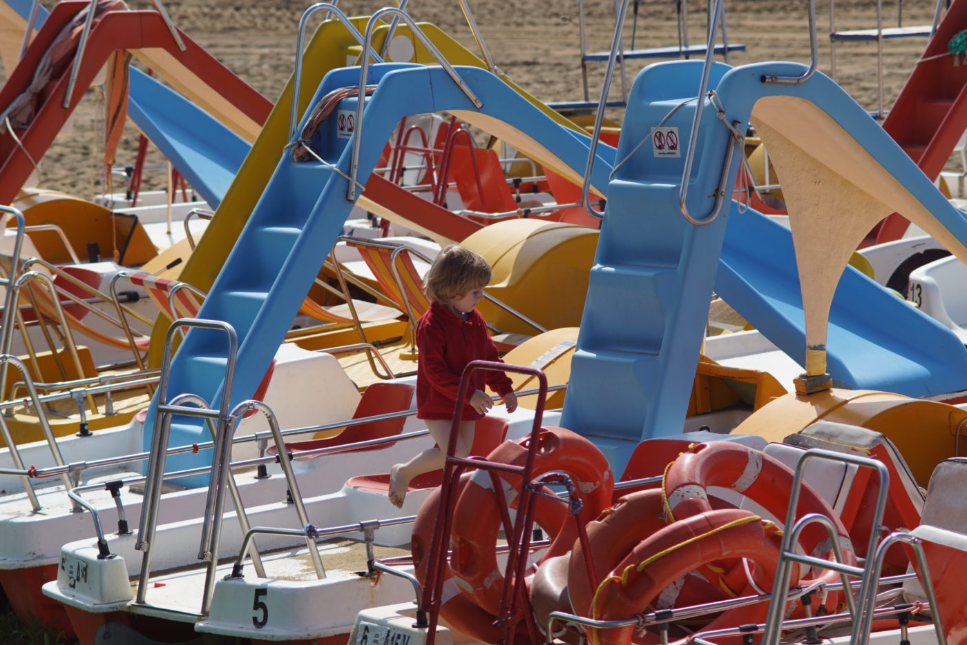 Es sind ca. zehn Tretboote mit Rutschen zu sehen die auf Sand am Strand liegen. Unser Sohn Luk hüpft in einem roten Langshirt von einem Boot zum anderen.