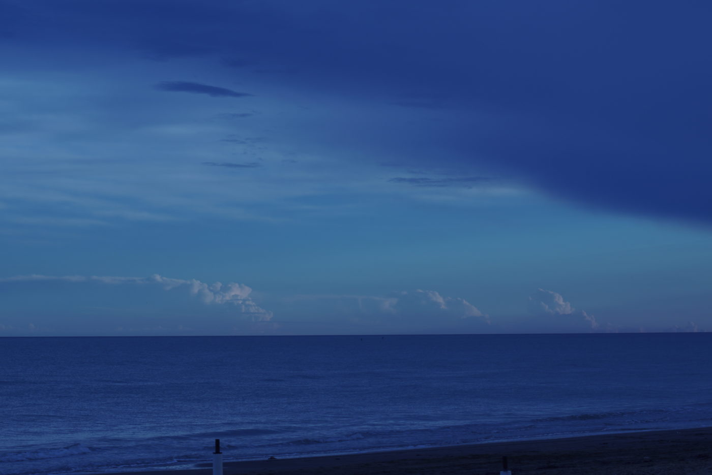 Das bild ist von vollen Blautönen geprägt. Im Vordergrund ist einstück Strand zu sehen und dahinter schließt die fast spiegelglatte Adria an. In der Ferne ist eine mächtige Wolkenbank zu sehen.