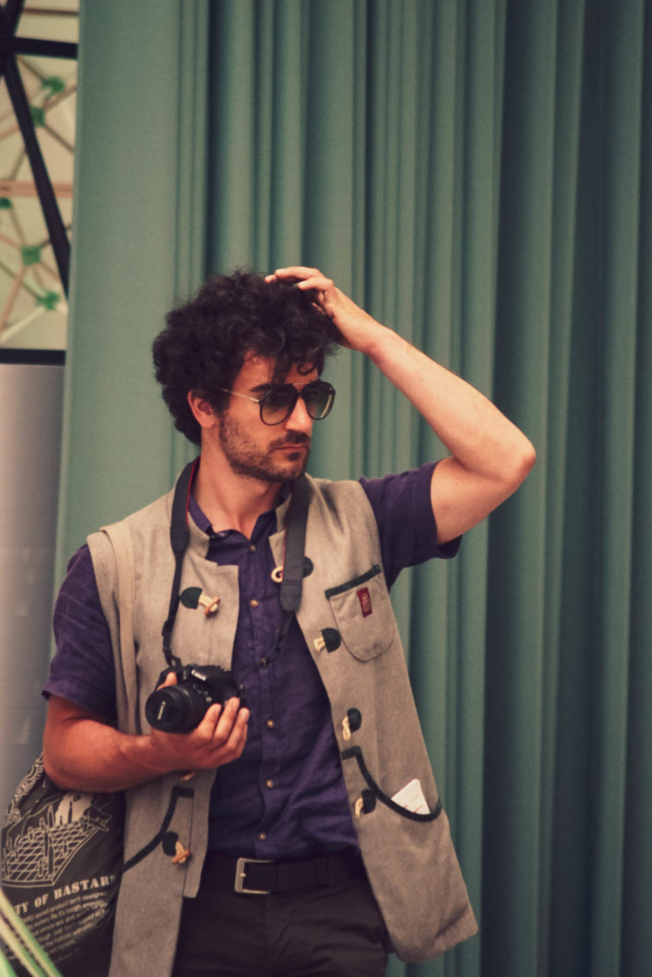 Ein Mann mit lila Hemd und beiger bayrischer Weste. Er hält eine Kamera in derr einen Hand und fährt sich mit der anderen Hand durch sein kurzes lockiges Haar