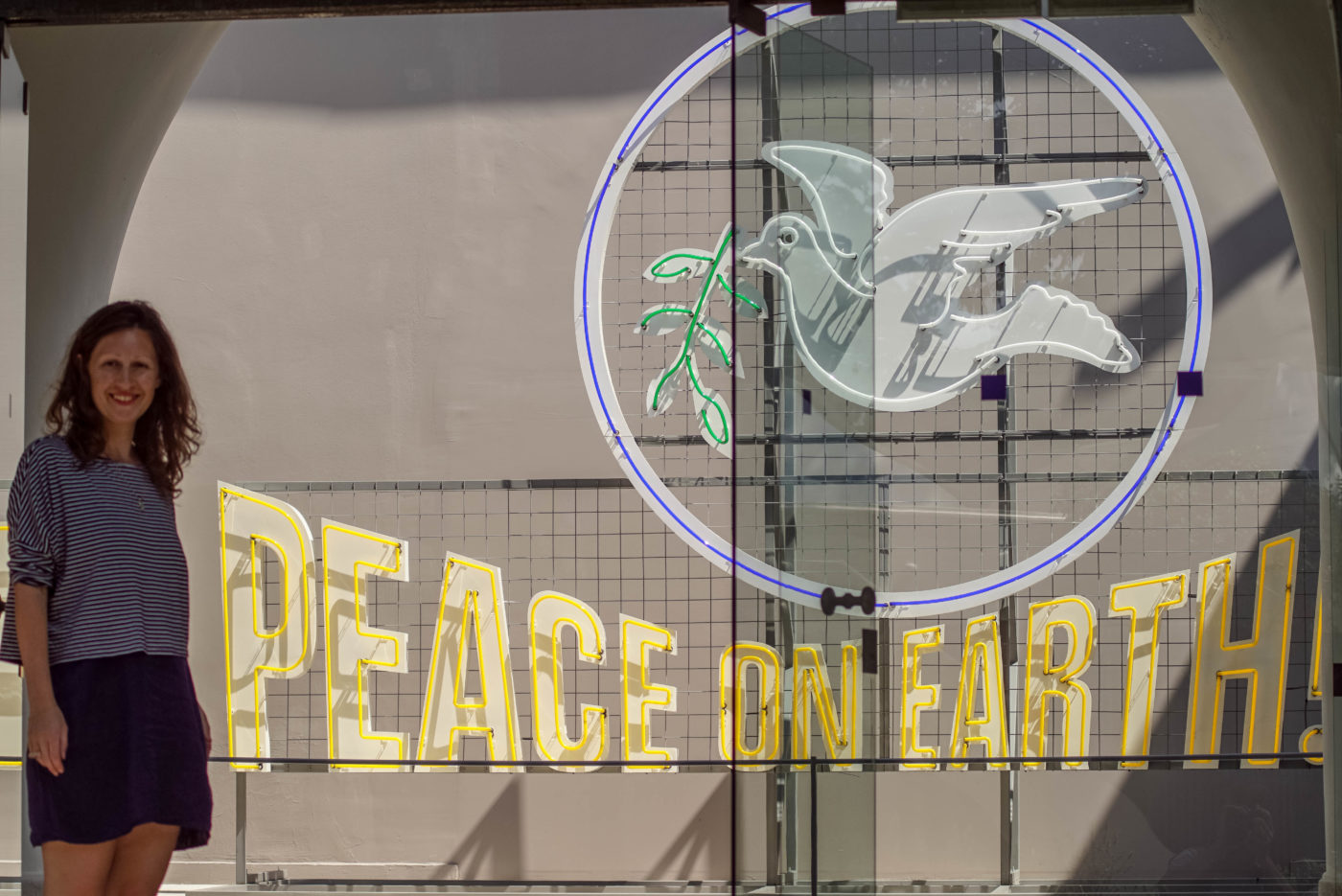 Lea steht vor dem ungarischen Pavillon Eingang und lächelt frölich in die Kamera. Neben ihr ist aus dünnen Neonröhren eine weiße Taube mit grünem zweig zu sehen, sie umschließt ein gelber Kreis. Darunter ist der Schriftzug "peace on earth!" zu lesen