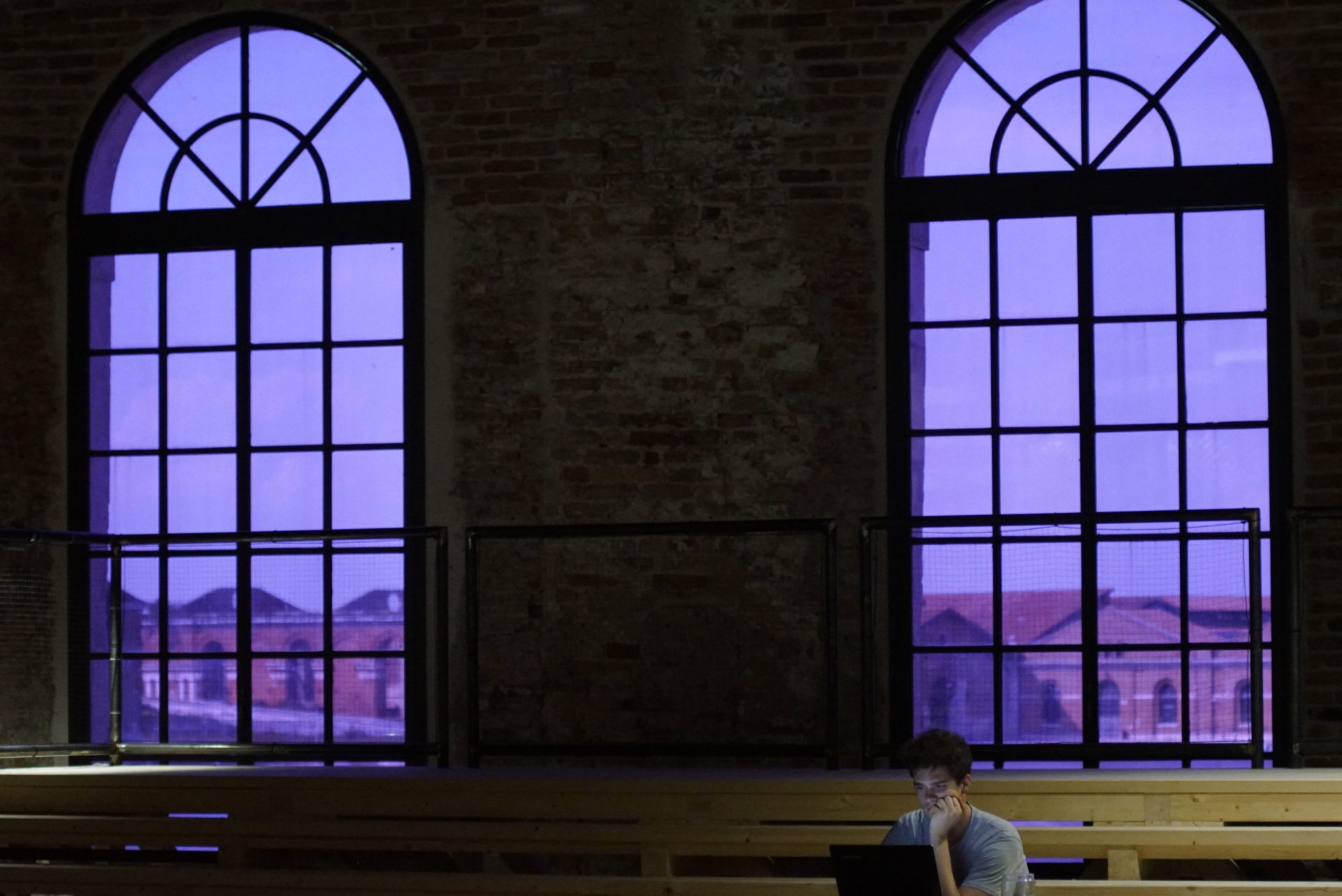 Zwei große lila Rundbogenfenster durch die man Teile des Arsenals von Venedig sieht. Im Vordeergrund sind lange helle Tische zu sehen an denen ein Mann in einen Laptop Blickt