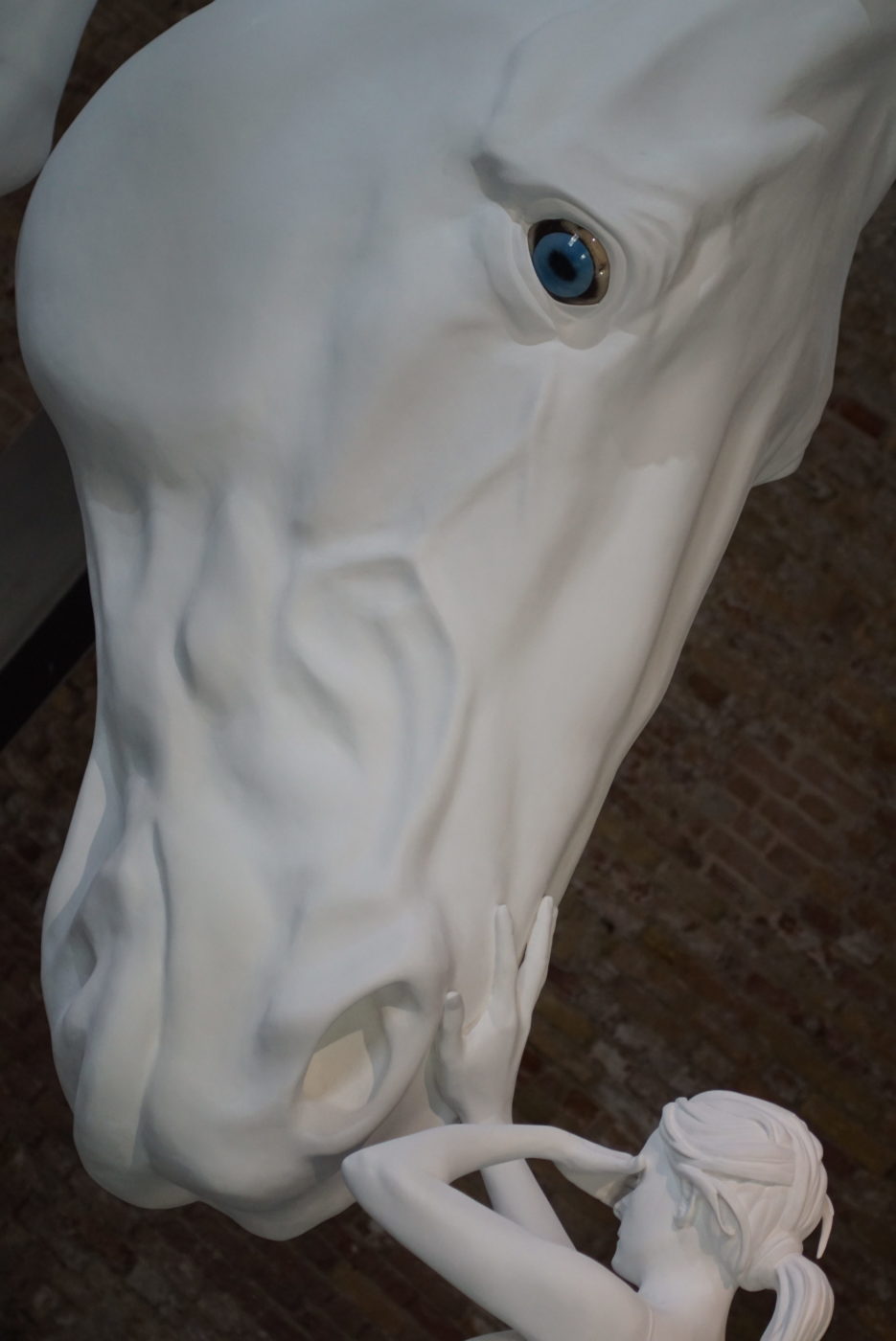 Überdemensionierter Pferdekopf aus Porzellan mit baluen Augen. Eine Frau aus Porzellan die so groß ist wie der Pferdekopf streichelt diesem die Nüstern und bedeckt sich mit der anderen Hand die Augen
